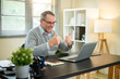 © bongkarn - Old white caucasian man wearing glasses laughing with thumbs up at laptop on office's working table.
