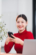 © PRIME STOCK LAB - Young Asian woman in red sweater smiling while using smartphone at desk, modern workspace, natural light, cheerful mood