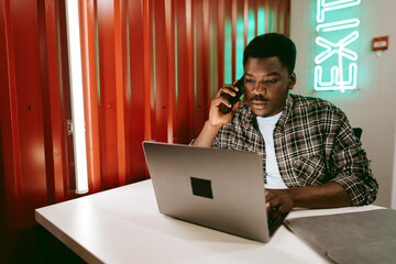 Poster - Young man working on laptop while on phone in a modern workspace with exit sign glowing