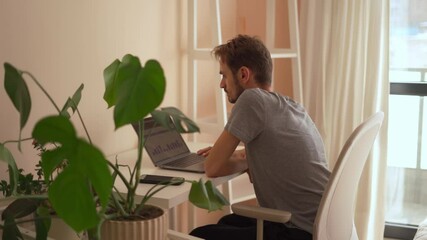 Poster - A young man works intently on a sleek laptop in a sunlit, beautifully designed workspace with vibrant plants, showcasing a blend of productivity and a serene, creative environment