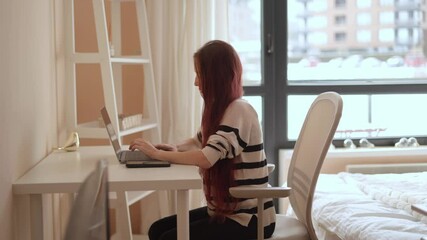 Poster - A young woman with long red hair sits at a contemporary desk, concentrating on her laptop. The bright, airy room features a cozy atmosphere with natural light