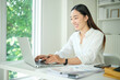 © wattana - Smiling Asian woman in a white shirt working on a laptop at a bright modern desk near a window
