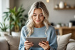 © f_bossa - A young woman with blonde hair smiles gently while looking at a tablet in a cozy living room setting.