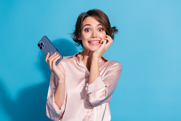 Poster - Young woman in pink satin blouse smiling joyfully, holding a smartphone against a vibrant blue background