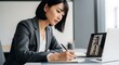 © Richafuji - Businesswoman taking notes during online meeting sitting at desk in modern office. Woman is wearing grey suit and gold necklace. Video call on laptop with blurred background.