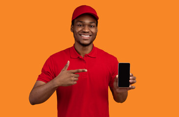 Poster - Choose Our Delivery Service. Smiling african american courier holding mobile phone with blank screen for mockup, wearing red cap and uniform, pointing at black empty gadget, orange studio background