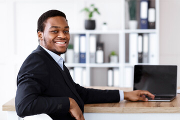 Successful african american businessman posing in his office, using modern laptop with blank screen, smiling at camera, copy space. Cheerful black manager in suit working on computer, mockup