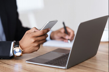 Aufkleber - Unrecognizable black manager in suit holding smartphone, taking notes, sitting in front of laptop, office interior, cropped. African american businessman using modern gadgets for business