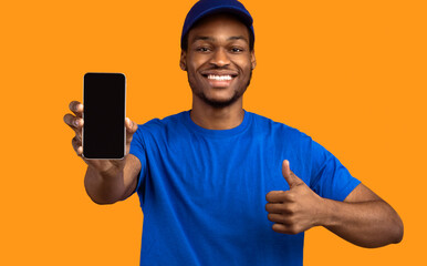Poster - Recommending Delivery Service. Smiling african american male courier holding smartphone with blank empty screen for mockup and showing thumbs up gesture, wearing blue cap and uniform, orange studio