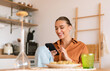 © Prostock-studio - Excited lady using smartphone in kitchen while having dinner, sitting at table and texting on phone, browsing social networks or shopping online, free space