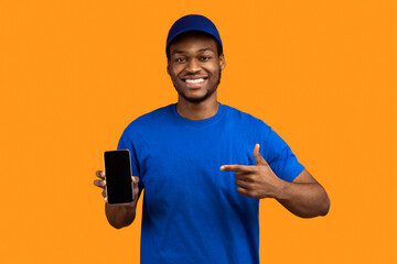 Poster - Recommending Service. Smiling african american delivery guy holding smartphone with blank screen for mockup, wearing blue cap and uniform, pointing at black empty monitor isolated on orange background