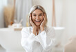 © Prostock-studio - Attractive blonde adult woman in bathrobe touching her face and smiling at camera, enjoying her daily face care routine, bathroom interior, copy space. Anti-age, lifting concept
