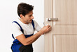 © Prostock-studio - Repair Service Concept. Portrait of smiling handsome young locksmith in blue uniform installing door knob on front wooden entrance door at home or office workplace, isolated on white studio background