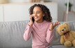 © Prostock-studio - Communication concept. Happy african american girl talking on smartphone, enjoying conversation with friend. Teen sitting on sofa with teddy bear nearby