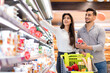 © Prostock-studio - Happy Arabic Spouses Buying Food And Dairy Products During Grocery Shopping Holding Milk Standing Near Shelf In Modern Supermarket. Happy Customers Purchasing Essentials In Store