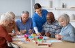 © Prostock-studio - Cheerful creative multiracial senior men and women sitting around table and drawing at nursing home, doing arts and crafts together, young lady nurse assisting group of elderly people, copy space