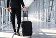 © Prostock-studio - Unrecognizable entrepreneur in black suit walking by airport terminal, carrying luggage and passport with flight tickets, copy space. Rich businessman with suitcase going to business trip, cropped