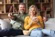 © Prostock-studio - Home Relax. Happy Married Middle Aged Couple Watching Tv In Living Room, Smiling Spouses Eating Popcorn And Drinking Coffee Together While Relaxing On Comfortable Couch, Closeup Shot