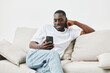 © SHOTPRIME STUDIO - Smiling young man relaxing on a couch while browsing on his smartphone, showcasing a casual lifestyle in a minimalist interior design
