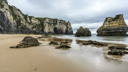 Poster - A dramatic coastal scene with rugged cliffs, rocky outcrops, and wet sandy beach under a cloudy sky.