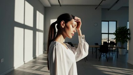 Poster - Professional businesswoman experiencing headache, pressing fingertips against forehead, standing near large windows with bright sunlight filling contemporary workspace