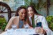 © carlesmiro - Two women, mother and daughter, are sitting at a table, drinking water and looking at a smartphone together, laughing and enjoying doing a videocall.