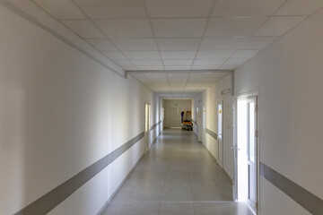  A long, empty hospital corridor with white walls, a grey stripe, and multiple doors. The sterile hallway features a tiled floor, and at the end of the corridor, a hospital bed with a catheter stand