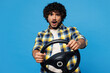 © ViDi Studio - Young sad shocked mad Indian man he wears yellow checkered shirt casual clothes hold steering wheel driving car look camera isolated on plain blue cyan background studio portrait. Lifestyle concept.