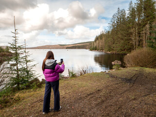 Wall Mural - Girl tourist enjoys view on a lake with cloudy sky reflection. Travel and tourism concept. Water is calm and green bushes and trees growing on the lake bank. Calm relaxed mood. Voyage and adventure.