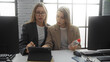 © Krakenimages.com - Women in modern office discussing work on digital tablet surrounded by computers, wearing suits, collaborating as business partners in a bright indoor workplace setting.
