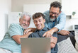 © luciano - Happy multigenerational family sitting on sofa using laptop together - grandfather with mature son and young grandson enjoying carefree moments together learning use of computer