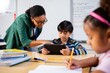 © Tom - Teacher assisting young boy with tablet in classroom while girl writes with pencil in foreground, focused and engaged learning environment