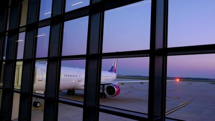 Poster - Modern passenger airplane parked on the airport runway viewed through the terminal window at dusk, with a beautiful purple sky in the background