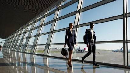 Poster - Professional businesspeople striding purposefully across modern airport terminal, passing expansive windows highlighting runway and parked aircraft, representing corporate travel dynamics