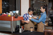 © yuttapon - Asian young woman adjusting apron for senior woman barista at coffee counter inside cafe showing care support and teamwork in small business with warm moment of generational connection and help