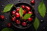 Top view of fresh sweet cherries in a bowl with water droplets on a stone background surrounded by leaves