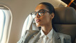 © Kowit - A professional woman in glasses sits in an airplane seat, gazing thoughtfully out the window during a flight.