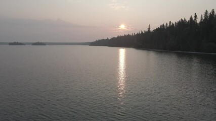 Wall Mural - Drone based aerial flying low above a large northern lake shrouded by forest fire smoke. The sun is rising above the spruce trees lining the shoreline.