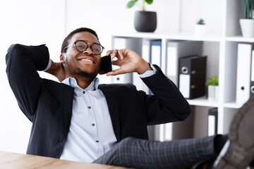 Poster - Relaxed black manager sitting with legs on workdesk, having conversation on mobile phone and smiling, talking to his lover while having break at work, resting after successful deal, free space