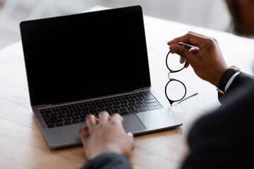 Aufkleber - Unrecognizable black businessman using laptop with blank screen, holding glasses, shot over shoulder, cropped, mockup. African american manager working online or attending webinar