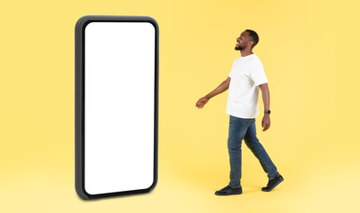 Aufkleber - Black Guy Walking Near Huge Phone With Empty Screen In Studio Over Yellow Background. Great Mobile Application Advertisement. Panorama, Full Length Shot