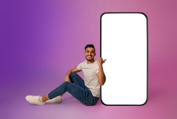 Poster - Full length of cheerful young Arab man sitting near big cellphone with mockup for mobile app on screen in neon light. Smartphone display template for new website or online ad
