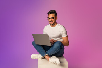 Poster - Full length of young Arab man sitting cross legged with laptop, having video call, working online in neon light. Happy middle Eastern guy watching movie, chatting on internet