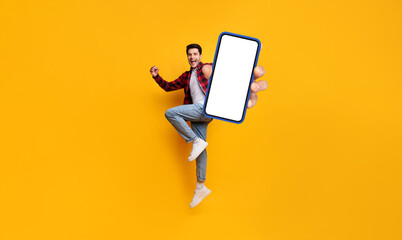 Poster - Yes. Full body length portrait of joyful man jumping up, making winner gesture with clenched fist and showing cell phone with free empty space for mobile app on white screen, yellow studio background