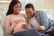 © Prostock-studio - Positive black man touching his pregnant girlfriend's tummy, communicating with baby, feeling its movements at home. African American couple hugging on sofa, waiting for new child