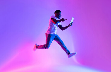 Poster - Excited young black guy with tablet computer jumping in neon light, copy space. Emotional African American man using modern touch pad and flying in air. Cool app or advertisement concept