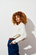 © SHOTPRIME STUDIO - Fashionable young woman with curly hair wearing a white sweater against a plain background, highlighting an elegant and modern aesthetic in casual attire