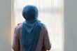 © oio - Woman wearing a blue hijab and light pink shirt standing quietly and gazing out a bright window with soft curtains, conveying a serene and contemplative mood