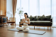 © Monster Ztudio - Young Asian woman meditating in lotus position at home, sitting on the floor in fitness clothing. Breathing workout to achieve relaxation, peace and mindfulness