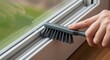 © velikiyzayats - Detailed close-up of a person's hand using a small cleaning brush to remove dirt and dust from a window frame track. Home improvement and household chore concept.